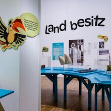 Foto in den Ausstellungsraum „Pippis Papa“. Im Vordergrund der Manu-Vogel (fiktiver Charakter). Im Hintergrund der groß geschriebene Titel eines Kapitels „Landbesitz“. Unter dieser Schrift ein vergrößertes Bild von Carl Petersen und seiner Frau Singdu.