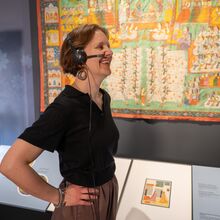 Foto eines Ausstellungsbereichs. Unter einem bunten antiken Wandteppich mit Alltagssituationen stehen Informationstafeln. links daneben eine Vitrine mit einem Exponat. Davor steht eine lächelnde Frau mit Headset, die ihre Hände in die Hüften stemmt.