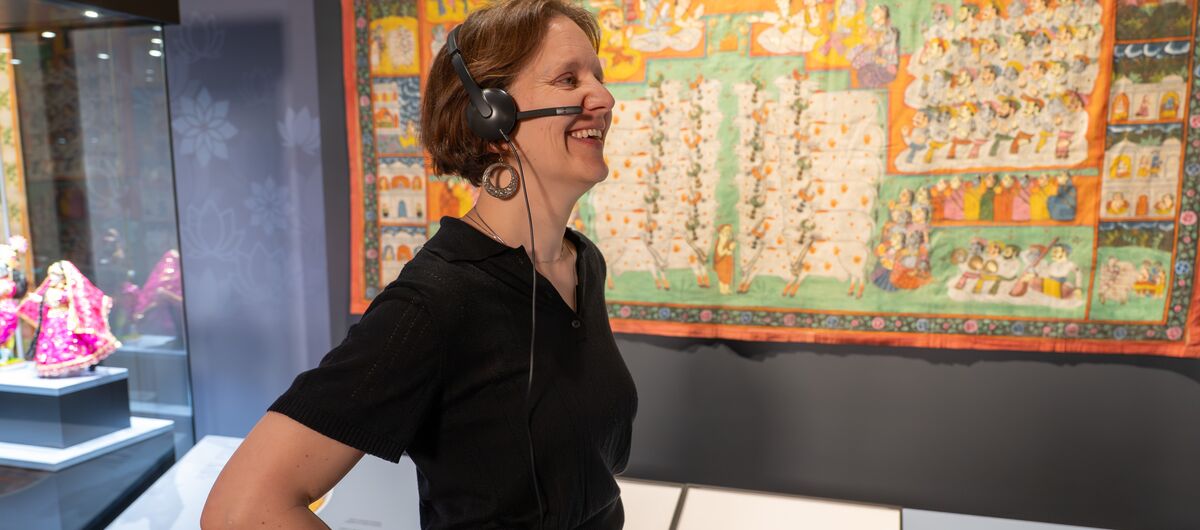 Foto eines Ausstellungsbereichs. Unter einem bunten antiken Wandteppich mit Alltagssituationen stehen Informationstafeln. links daneben eine Vitrine mit einem Exponat. Davor steht eine lächelnde Frau mit Headset, die ihre Hände in die Hüften stemmt.