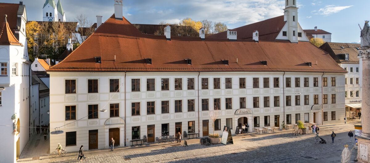 Foto eines breiten dreigeschossigen Hauses mit Ziegelrotem Dach inmitten einer Stadt. In der Breite zieren jeweils 21 Fenster die Front des 1. und 2. Geschosses. Die Front des Erdgeschosses wird ähnlich symmetrisch durch Fenster und Türen geschmückt.