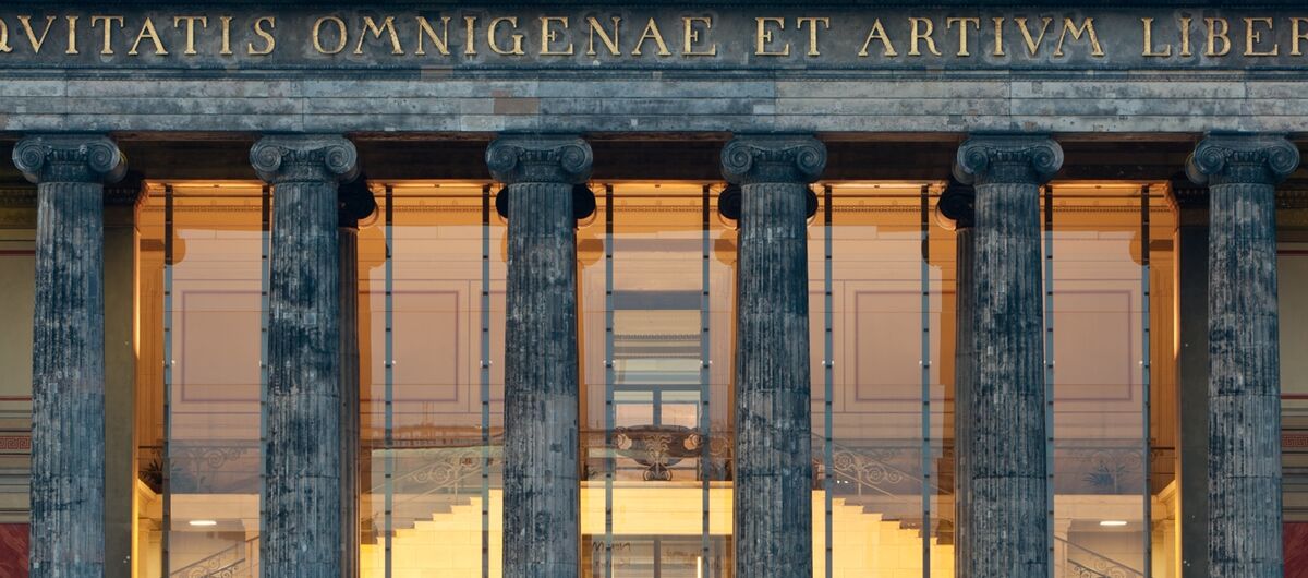 Foto des Eingangsbereichs des Alten Museums in Berlin. Treppen führen hoch zu einer großen Glasfront mit Glastür, die durch sechs pompöse Säulen geschützt wird. Darüber eine lateinische Inschrift in goldenen Buchstaben. Im Inneren ist ein warmes Licht.