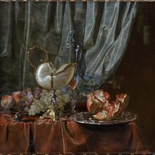 Stillleben mit Nautiluspokal, Pfirsichen, dunklen und grünen Trauben sowie einem auseinandergenommenem Granatapfel. auf einem Silbertablett. Das Stelleben steht auf einem roten Tuch. Dahinter hängt ein grauer Vorhang.