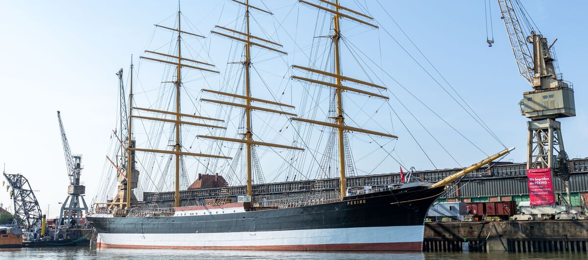Der Frachtsegler Peking liegt im Hafen vor Anker. Es ist ein vier Masten tragendes Schiff mit insgesamt 32 Segeln, die eingeholt sind. Rechts und links des Schiffes steht je ein Hafenkran. Es ist ein freundlicher Tag.