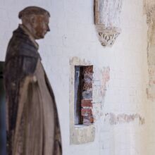 Foto in schräger Perspektive auf eine weiß gestrichene Backsteinwand., in die ein kleiner Durchbruch geschlagen wurde. Im Vordergrund steht eine Dominikanerstatue.