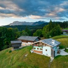 Luftaufnahme eines Bereichs des Freilichtmuseums Glentleiten. Traditionelle Hofgebäude stehen auf einem Berg. Auf der Abschüssigen Wiese unter den Gebäuden stehen weiße und schwarze Schafe. Im Hintergrund sind Wald und Berge zu sehen.