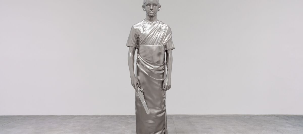 Foto einer Statue, die vor einer weißen Wand und auf grauem Betonboden steht. Sie ist silbern. Dargestellt wird ein geradestehender Mensch mit kurzen Haaren, knöchellanger Tunika und Schwert in seiner rechten Hand. Der Gesichtsausdruck ist neutral.