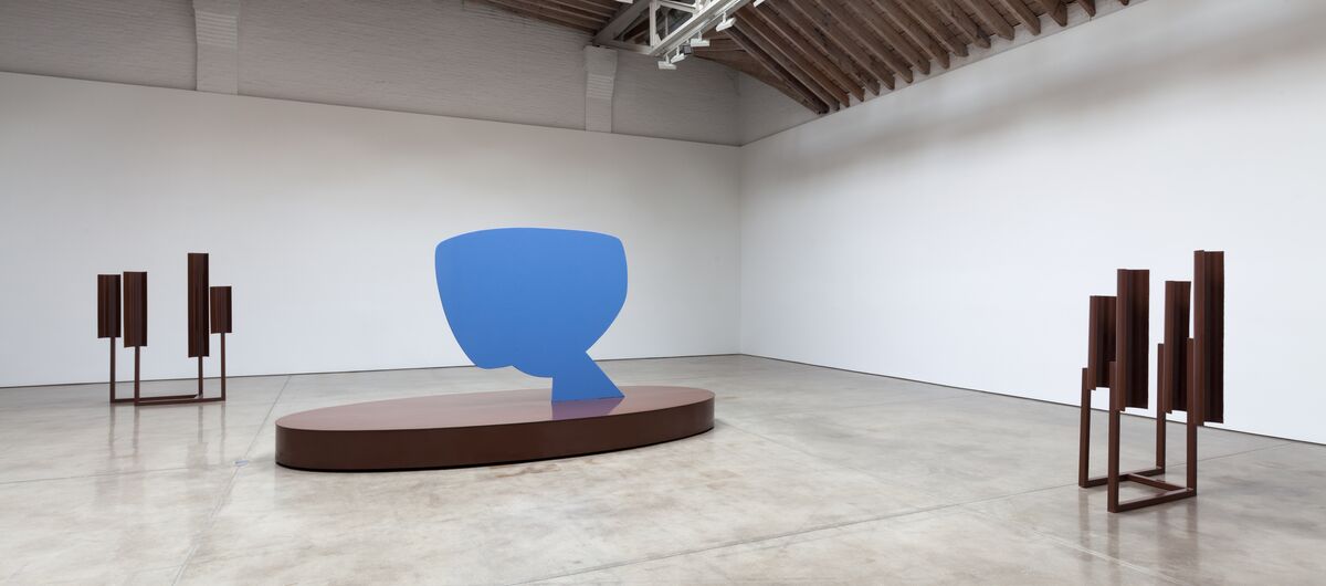 Foto eines großen minimalistischen Ausstellungsraums. Im Raum stehen drei abstrakte Skulpturen. Die linke und rechte Skulptur sind braun. Die in der Mitte besteht aus einem braunen ovalen Sockel, auf dem senkrecht eine blaue sprechblasenartige Platte ist.