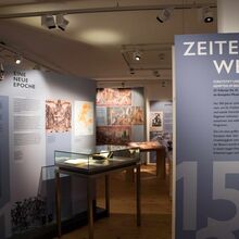 Foto eines Teils der Ausstellung Zeitenwende. In einem dunklen Raum stehen in blau gehaltene Informationswände sowie Vitrinen, die durch Lichtspotts angestrahlt werden. Auf eine Wand wird etwas projiziert.