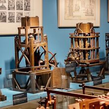 Foto der Modellkammer des Maximilianmuseums. Zu sehen sind fünf verschiedene Maschinen, eine weitere steht im Vordergrund. An der Wand dahinter hängen vier Bilder mit Skizzen und Karten.