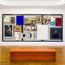 Foto eines Ausstellungsbereichs. Im Vordergrund steht eine einfache orangene Bank. Dahinter vor einer Wand steht auf einem weißen Sockel ein großes schwarzes dreigeteiltes Gitter. An diesem hängen Gemälde unterschiedlichster Stile, Größen und Formen.