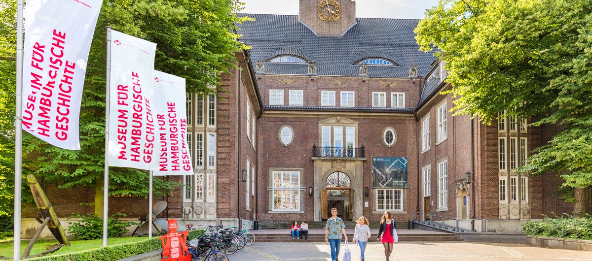 Menschen vor einem palastähnlichen Gebäude. Der rote Backsteinbau geht über 5 Etagen. Der Eingangsbereich ist über Treppen erreichbar, hat ein steinernes Tor und ist von Fenstern umrandet. Darüber ein Balkon. Auf dem Dach ist mittig ein Turm.