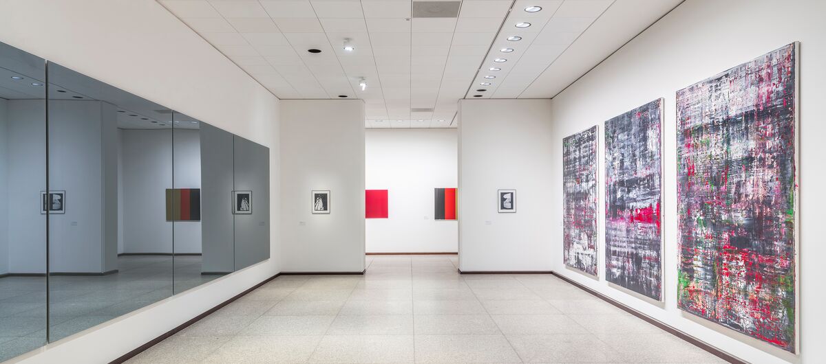 Ausstellungsansicht „Gerhard Richter. 100 Werke für Berlin“. In einem langen Raum hängen an der rechten weißen Wand drei große abstrakte Gemälde. Gegenüber auf der linken Seite hängen vier dunkle Spiegel. Geradeaus ist ein Durchgang zu einem weiteren Raum