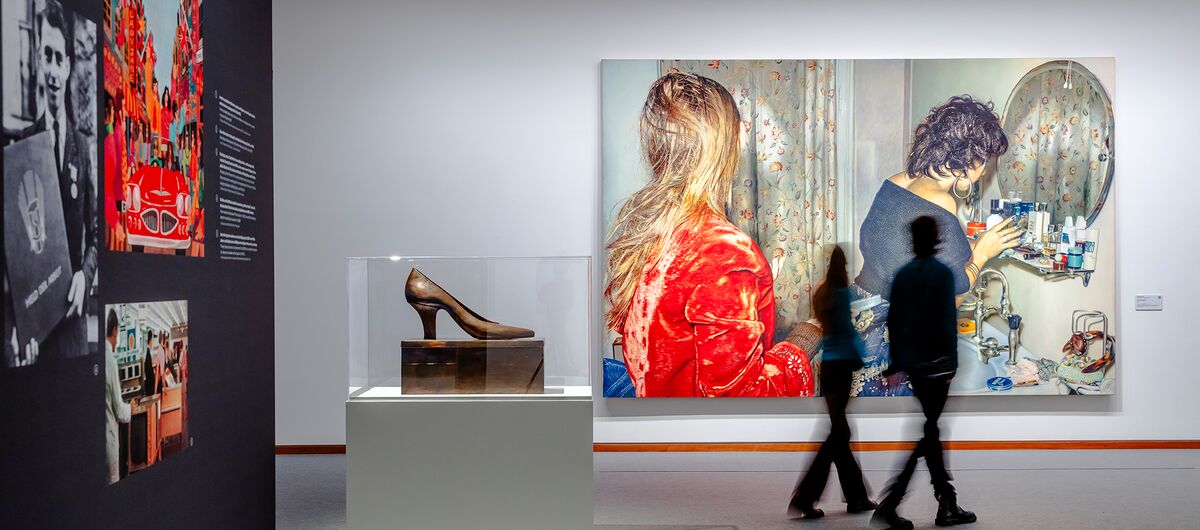Foto der Ausstellungsansichten zu „Zerreißprobe. Kunst zwischen Politik und Gesellschaft. Sammlung der Nationalgalerie 1945-2000“. In einer Vitrine steht ein großer Damenschuh auf einem Sockel. Dahinter hängt ein großes Foto von zwei Frauen im Bad.