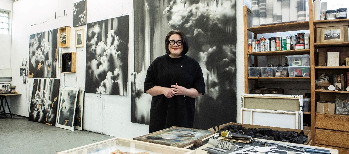 Foto der Künstlerin Lucia Tallová im Atelier. Sie steht hinter einem großen Arbeitstisch und lacht. Rechts hinter ihr ist ein Holzregal mit Kunstmaterialien. Links hinter ihr hängen und stehen schwarz-weiß Bilder in unterschiedlichen Größen.