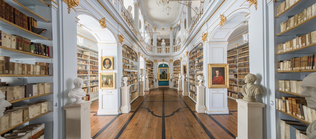 Historischer Bibliothekssaal mit breitem Holzgang unter kunstvoll verzierten Deckengewölbe. Die Wände zieren Regale mit goldenen Elementen, die Bücher, Porträtgemälden und Büsten enthalten.