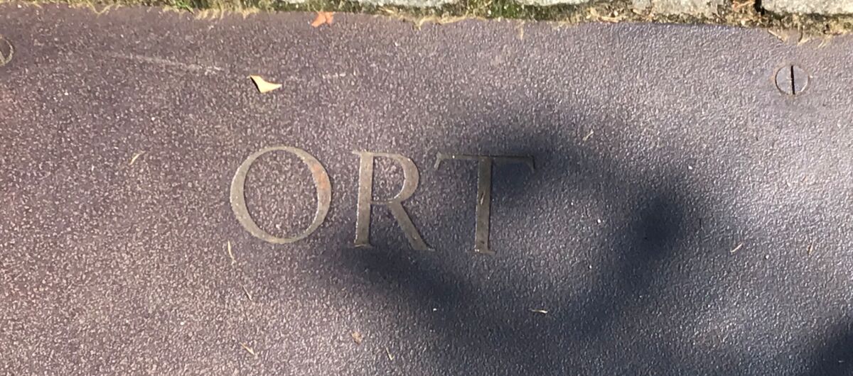 Schriftzug "Ort" eingraviert auf eine Stahlplatte, die an kleine graue Steinquadrate grenzt, welche den Bodenbelag bilden. Durch die Ritzen der Steine dringt grünes Moos.