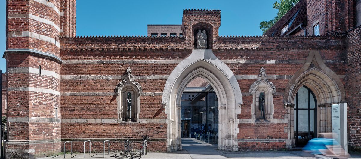 Foto von St. Annen bei Sonnenschein. Der Backsteinbau ist durch waagerechte Sandsteinlinien durchzogen. Die Mauer ist durch einen Durchgang unterbrochen. Rechts und links daneben ist je eine Einbuchtung mit einer Figur zu sehen.