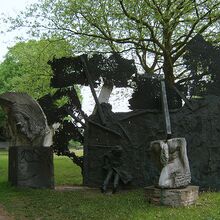 Foto eines Denkmals für den 2. Weltkrieg, das aus drei Teilen besteht. Zwei kleinere abstrakte Elemente aus Stein stehen je auf einem Steinsockel. Das größte Element ist schwarz, flächig und eine Person ragt mit Beinen und Mantel heraus, ebenso Stangen.