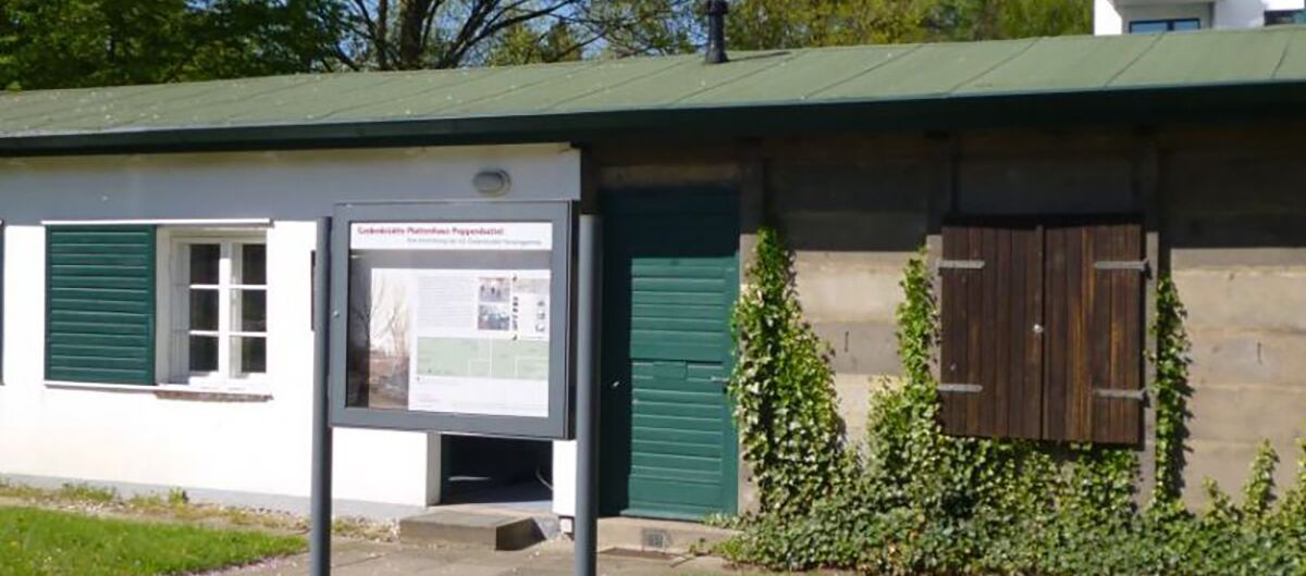 Langes, flaches Gebäude, auf der einen Seite weiß gestrichen mit grünen Fensterläden und auf der anderen Seite grau/braun mit dunkelbraunem, geschlossenen Fensterladen
