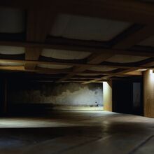 Foto in einen Raum ohne Einrichtungsgestände und Tapeten. An der Decke sind breite Holzbalken schachbrettartig angeordnet. Drei Holzbretterverschläge sind an den Seiten, an denen je eine brennende Lampe herunterstrahlt. Ansonsten ist es dunkel.