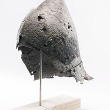 Foto einer historischen Beckenhaube. Sie ist auf einen Stab gesetzt, der in einen hellen Holzsockel mündet. Der eiserne Helm weist eine unebene Oberfläche auf. Teilweise Durchziehen ihn Löcher. Die Ränder sind ausgefranst.