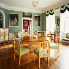 Foto eines barocken Zimmers mit Holzboden. Die Wände, an denen Gemälde hängen, sind weiß und grün gestrichen. In der Mitte steht ein runder Holztisch mit sieben Holzstühlen, deren Sitzflächen grün-weiß gestreift bezogen sind.