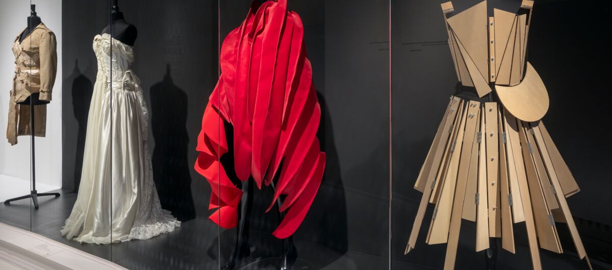 Ausstellung von verschiedenen Kleidungsstücken. Ein Kleid ist wie ein X geschneidert und wirkt wie aus Holzstreben. Dahinter ein rotes, bauschiges Kleid, wie ein Koralle. Dahinter ein Brautkleid-ähnliches Stück sowie ein Oberteil mit schrägem Seitenteil.