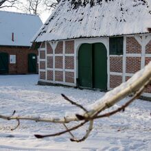 Foto eines Bauernhauses mit Strohdach. Seitlich dahinter steht ein Schuppen bzw. Stall, mit Dachpfannen gedeckt. Auf dem Hof und auf den Dächern liegt Schnee. Im Vordergrund ragt ein mit Schnee bedeckter Ast in das Foto.