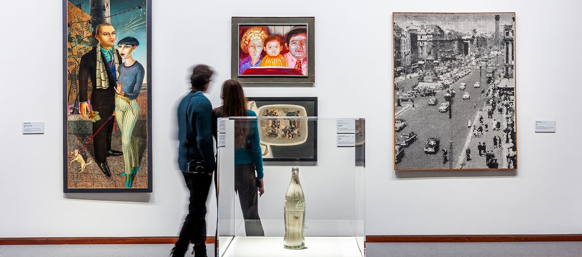 Foto eines Ausstellungsbereichs. Im Vordergrund mittig steht in einer Vitrine eine metallisch wirkende Coca-Cola-Flasche. Dahinter sind vier Gemälde unterschiedlicher Form und Stil, die von zwei Menschen betrachtet werden.