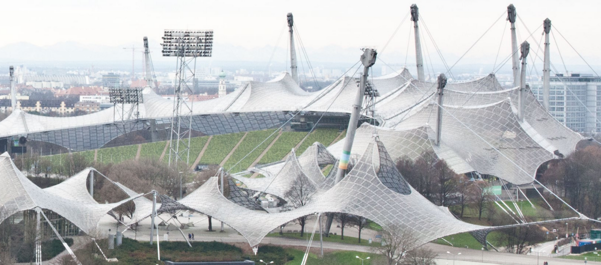 Foto der Zeltdachkonstruktion des Münchener Olympiastadions. Wie Spinnennetze sind die weiß durchsichtigen Dächer über die relevanten Areale gespannt.