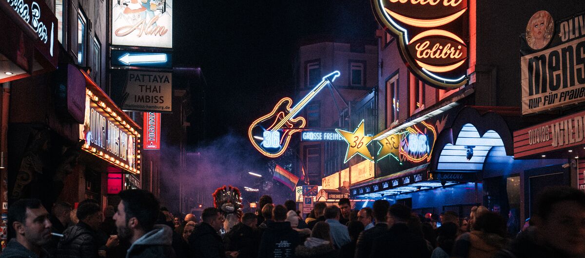 Foto von der Reeperbahn. Es ist dunkel. Auf der Straße stehen Trauben mit Menschen. Rechts und links an den Häuserfassaden strahlen bunt Leuchtreklamen.