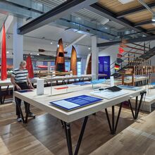 Foto eines Ausstellungsbereichs des Wassersportmuseums. Auf großen Tischen sind Informationsschilder, Paddel und Kanus ausgestellt.