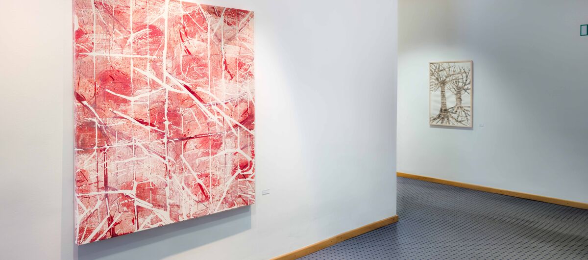 Foto eines Flures mit blauem Teppichboden und weißen Wänden. An der Wand im rechten Vordergrund hängt eine große quadratische Leinwand mit abstrakter Malerei mit roten, braunen und weißen Farben. Im Hintergrund hängt ein kleineres Bild mit zwei Bäumen.