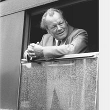 Schwarz-weiß Foto von Willy Brandt, der in einem Zug steht und sich mit seinen gefalteten Händen auf das heruntergelassene Fenster abstützt. Er schaut freundlich nach links. Das Fenster ist nass.