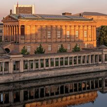 Foto der Museumsinsel Berlin. Im Vordergrund ist die Spree. Dahinter sind die Alte Nationalgalerie, Teile des Pergamonmuseums und des Neuen Museums sowie des Kolonnadenhofs zu sehen -  alles alte, prächtige und aus Stein errichtete Bauwerke.