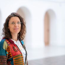 Foto in einem hellen schlichten Raum. Im Vordergrund ist auf der rechten Seite der Oberkörper eine Frau zu sehen. Sie schaut die Betrachtenden freundlich an. im Hintergrund sind Rundbogendurchgänge zu sehen.
