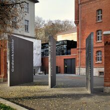 Foto des Eingangsbereichs der Gedenk- und Bildungsstätte Andreasstraße. Auf dem Vorplatz stehen senkrecht nebeneinander drei rechteckige schmale Stelen, auf denen senkrecht Worte stehen. Sichtbar ist das Wort "Gedenkstätte".