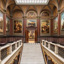 Foto des Treppenhauses in der Lichtwark-Galerie der Hamburger Kunsthalle. Eine Steintreppe, die in der unteren Bildmitte ist, führt in ein weiteres Geschoss mit historischen Bildern und Wandverzierungen.