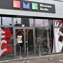 Foto des Eingangsbereichs zum Illuseum in Berlin. Die Glasfront ist mit großen Aufklebern beklebt, die z. B. abstrakte Hände abbilden, die einen Schatten in Form eines Bären werfen.