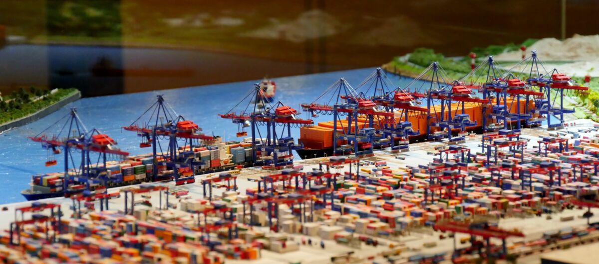 Foto eines Hafenmodells. Im Hintergrund ist Gewässer zu sehen. Davor liegen beladene Containerschiffe an der Kaimauer. Kräne und viele Container sind des Weiteren zu sehen.