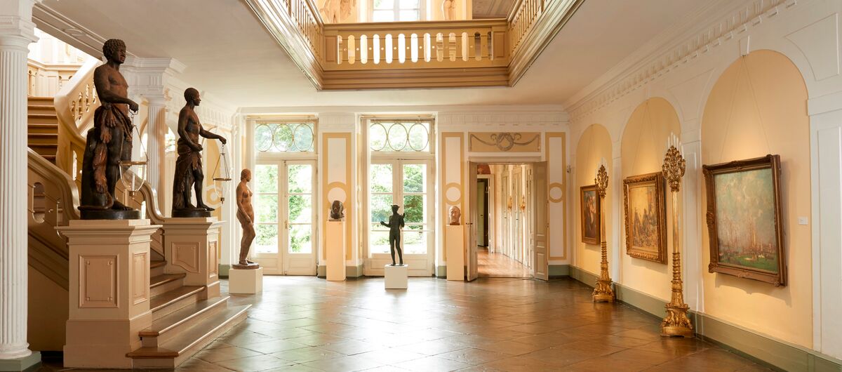 Foto der Diele des Behnhaus. Der Boden besteht aus alten großen speckigen Fliesen. Auf der linken Seite geht eine prachtvolle Treppe hoch, die am unteren Ende durch durch Statuen gesäumt ist. Auf der rechten Seite hängen drei große Gemälde an der Wand.