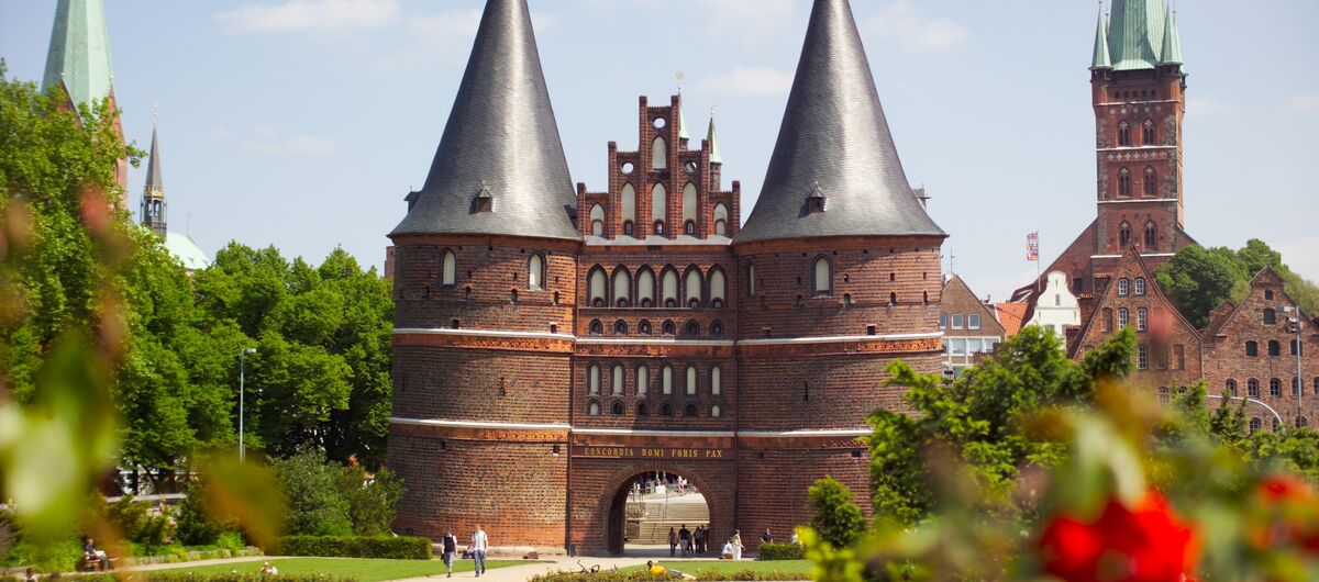 Foto des Holstentors bei Sonnenschein. Es ist ein Backsteinbau und besteht aus zwei großen Rundtürmen mit rundem spitzen Dach. Verbunden werden die Türme durch den Torbogen, über den sich weitere drei Geschosse ziehen. Davor ist eine Parkfläche mit Rasen.