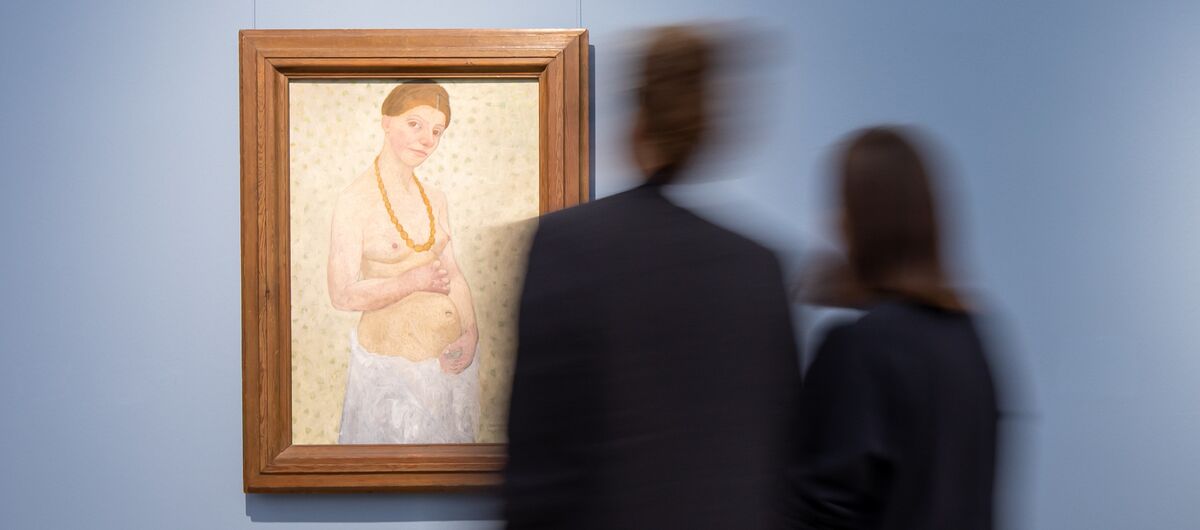 Foto zweier Menschen, die vor einem Gemälde stehen. Die Menschen sind unscharf, das Gemälde scharf. Auf dem Gemälde ist eine vermutlich schwangere Frau abgebildet, die nur eine große Perlenkette und ein weißes Laken um die Hüftenträgt.