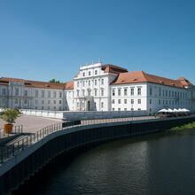 Foto des Schloss Oranienburg. Ein prächtiges weißes Gebäude mit rotem Dach steht im Zentrum des Fotos. Die rechte untere Ecke dominiert ein ruhiges Gewässer. Das Wetter ist sonnig.