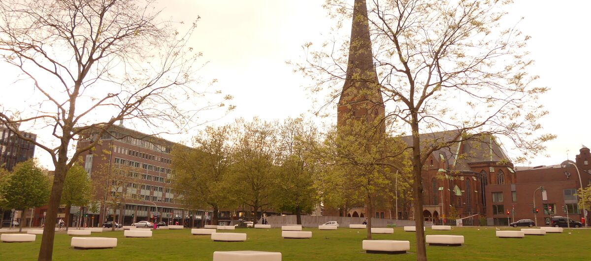 Das Foto zeigt eine große Wiese mit einzelnen Bäumen, auf der weiße quadratische Elemente als Bänke zur Verfügung stehen. Im Hintergrund sind moderne mehrgeschossige Geschäftshäuser und eine Kirche zu sehen.