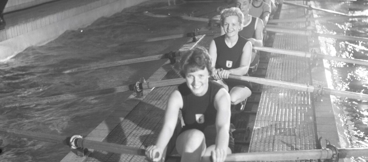 Schwarz-weiß Foto mit dem Titel „Versehrten-Rudern“, aufgenommen am 19.2.1962 in Berlin. In einem festinstallierte Rudertrainingsgerät mit sieben Plätzen sitzen in Sportkleidung freundlich schauende Frauen und Männer.