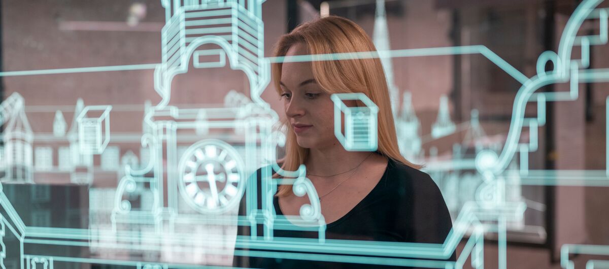 Foto eines Ausstellungsbereichs. Im Vordergrund steht eine Glasscheibe mit Aufdruck eines alten Gebäudes in weiß. Dahinter steht eine junge Frau mit schulterlangen blonden Haaren, die sich das Exponat anschaut.