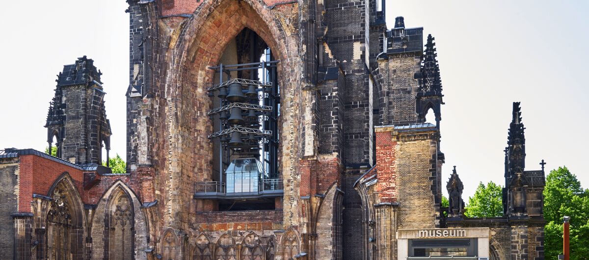 Frontaler Blick auf die Überreste von St. Nikolai mit Glocken im Kirchenschiff in rot-beigen Stein mit schwarzen Verfärbungen. Moderne Bauelemente, wie die Eingangstür zum Museum, fügen sich in den alten Bau ein.