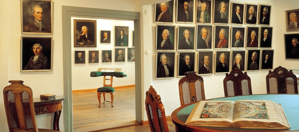 Blick in einen Raum, an den ein weiterer Raum durch eine geöffnete Tür anschließt. Sowohl im vorderen als auch im hinteren Raum hängen dicht aneinander Portraits von vielen Männern und wenigen Frauen aus dem 18. Jahrhundert.