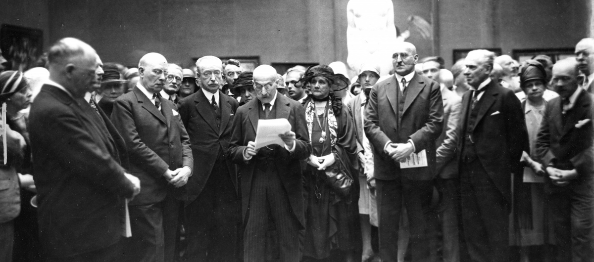 Schwarz-weiß Foto einer Gruppe von Männern und Frauen in einem Raum, die sich um einen Mann versammeln, der ein Blattpapier in den Händen hält und etwas vorzulesen scheint. Alle sind schick gekleidet und haben eine neutrale bis ernste Miene.